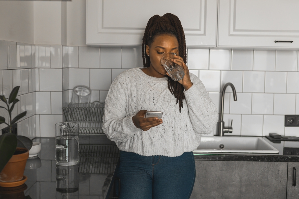 Portrait of African American female student dressed casually holding mobile phone and typing messages and communicating with friends via social networks using high-Internet connection in kitchen woman drinking a glass of water in her kitchen while looking at phone